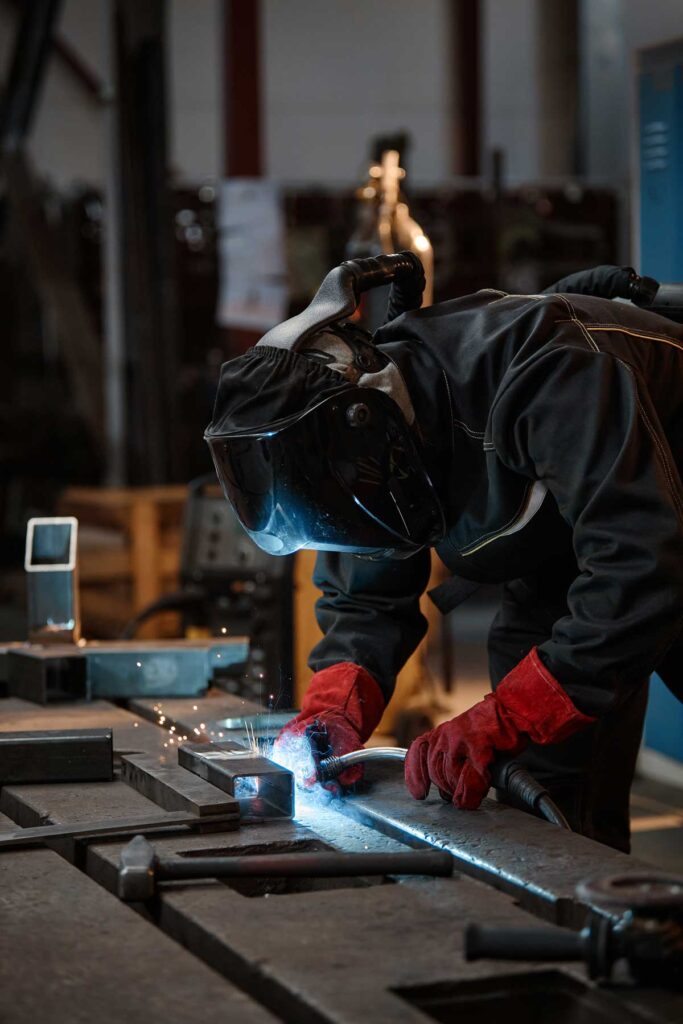 image of young adult welder in protective mask and gloves welding metal parts in workshop